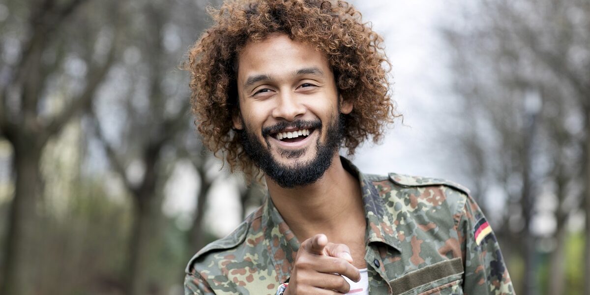 Portrait of smiling man with dyed ringlets wearing camouflage shirt pointing on viewer